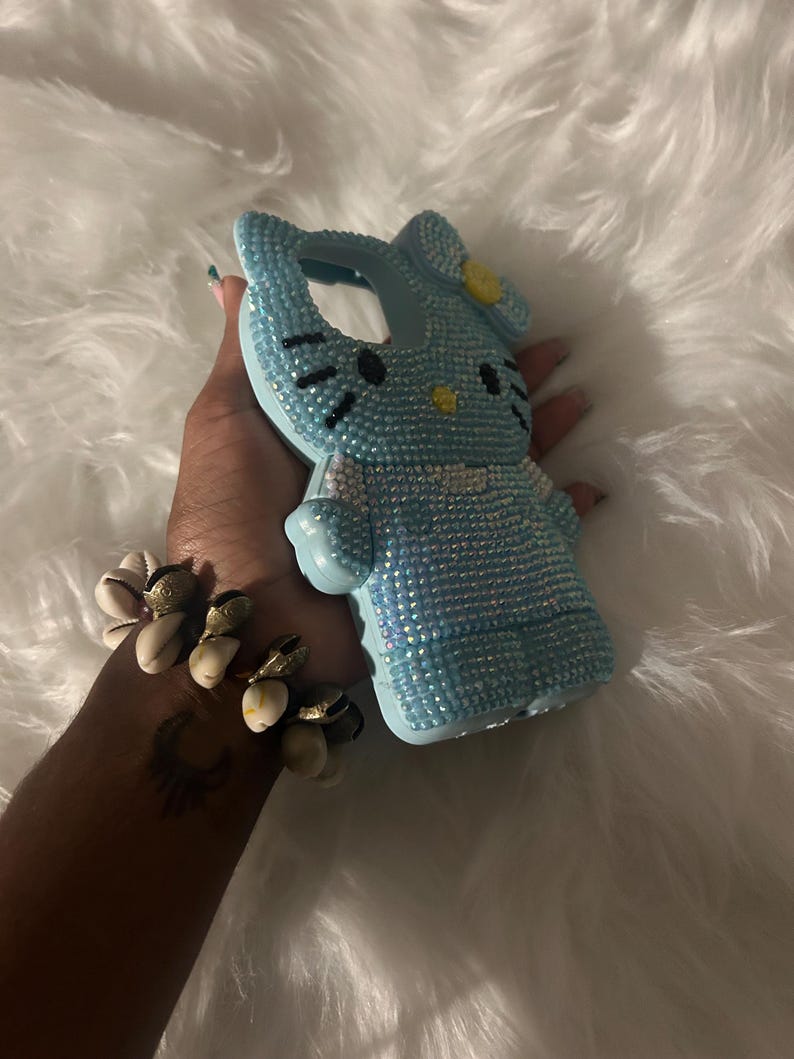K&ouml;nnte beinhalten: Eine hellblaue Handyh&uuml;lle in Form einer Zeichentrickkatze, bedeckt mit kleinen, glitzernden Strasssteinen. Die Katze hat schwarze Augen und Schnurrhaare sowie eine gelbe Schleife. Die H&uuml;lle befindet sich vor einem flauschigen wei&szlig;en Hintergrund.