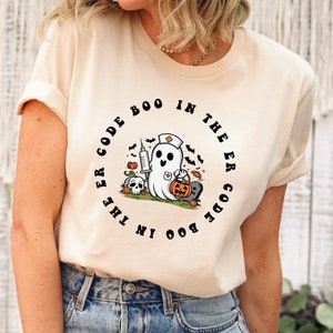 May include: A cream colored t-shirt with a black circle graphic featuring a ghost wearing a nurse's hat and holding a pumpkin. The text around the graphic reads "Boo in the ER Code Boo in the ER Code".