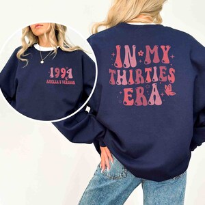 May include: A navy blue sweatshirt with a pink and red graphic on the back that says "In My Thirties Era" with a butterfly and stars. The front of the sweatshirt has a pink and red graphic that says "1994 Amelia's Version".