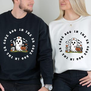 May include: Black and white sweatshirt with a ghost wearing a nurse's hat and holding a syringe and a pumpkin. The text on the sweatshirt reads "Code Boo in the ER Code Boo in the ER".