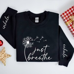 May include: Black sweatshirt with white text that reads "inhale" on the left sleeve, "just breathe" on the front with a dandelion graphic, and "exhale" on the right sleeve.