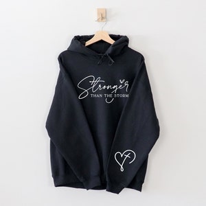 Pu&ograve; includere: Felpa con cappuccio nera con testo bianco che recita "Stronger than the storm" e un cuore bianco con una croce all'interno sul manico sinistro.