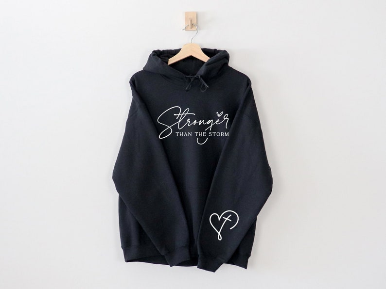 Pu&ograve; includere: Felpa con cappuccio nera con testo bianco che recita "Stronger than the storm" e un cuore bianco con una croce all'interno sul manico sinistro.