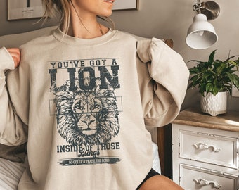 Può includere: Una felpa beige con una grafica di un leone e il testo "You've got a lion inside of those lungs, so get up & praise the Lord."