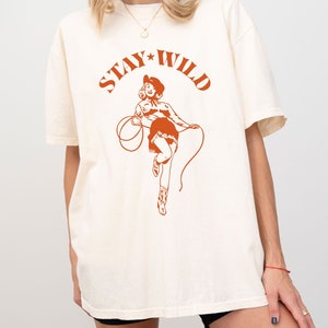 May include: A white t-shirt with a brown graphic of a woman in a cowboy hat holding a lasso. The graphic says "Stay Wild" with a star between the words.