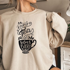 May include: A beige sweatshirt with a black graphic of a teacup and the text "A cup of tea solves everything".