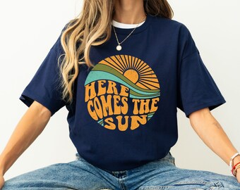 May include: A navy blue t-shirt with a retro-style graphic of a sun rising over a wave. The graphic includes the text "Here Comes The Sun" in a vintage font.