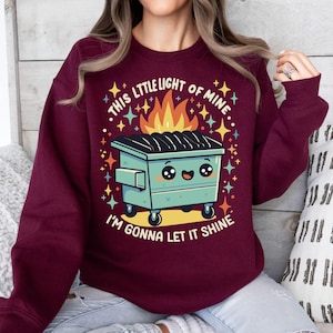 May include: A maroon sweatshirt with a graphic of a green dumpster with a smiling face and flames coming out of the top. The text "This little light of mine... I'm gonna let it shine" is printed on the sweatshirt.
