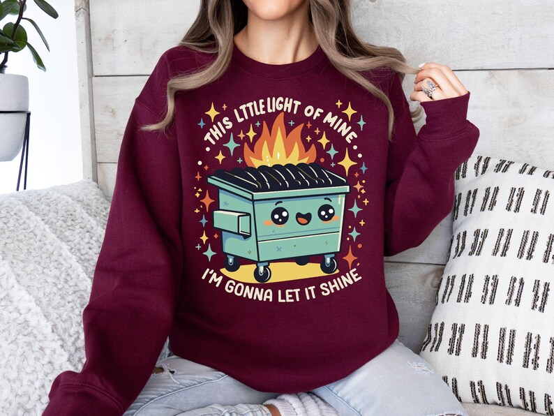 May include: A maroon sweatshirt with a graphic of a green dumpster with a smiling face and flames coming out of the top. The text "This little light of mine... I'm gonna let it shine" is printed on the sweatshirt.