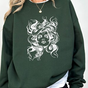 May include: A dark green sweatshirt with a white graphic print of Medusa's head with snakes for hair.