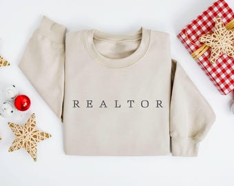 Op de afbeelding: Een beige sweater met ronde hals en het woord REALTOR in zwarte hoofdletters op het midden van de borst. Het kledingstuk heeft lange mouwen en een geribbelde halslijn, boorden en zoom, gepresenteerd in een platte lay-out.