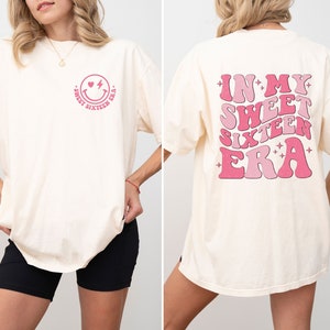 May include: A white t-shirt with a pink smiley face with a heart for an eye and a lightning bolt for a mouth. The text "Sweet Sixteen Era" is written around the smiley face. The back of the shirt has the text "In My Sweet Sixteen Era" written in pink with a retro font and stars.