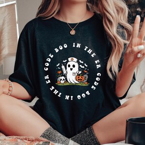 May include: Black t-shirt with a white circle graphic featuring a ghost wearing a nurse's hat and holding a syringe. The text around the ghost reads "Boo in the ER Code the ER Code Boo".