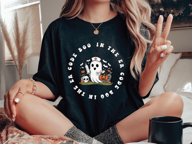 May include: Black t-shirt with a white circle graphic featuring a ghost wearing a nurse's hat and holding a syringe. The text around the ghost reads "Boo in the ER Code the ER Code Boo".
