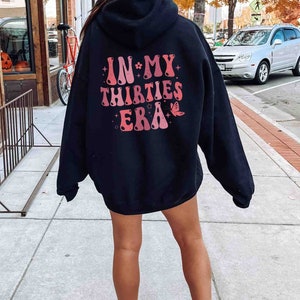 May include: Black hooded sweatshirt with a pink and red graphic print on the back that says "IN MY THIRTIES ERA" with a butterfly design.