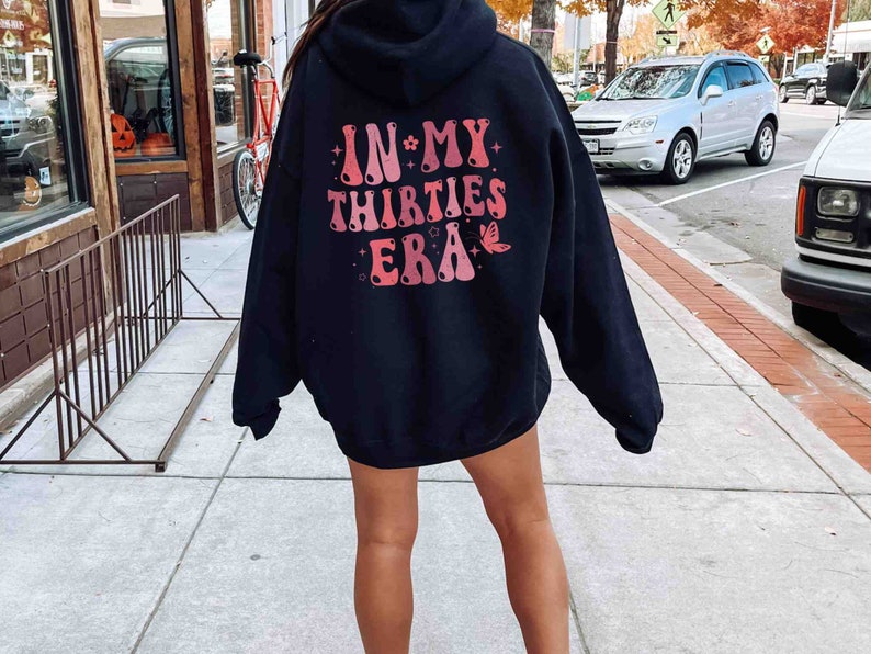 May include: Black hooded sweatshirt with a pink and red graphic print on the back that says "IN MY THIRTIES ERA" with a butterfly design.