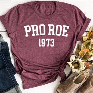 May include: A maroon t-shirt with white text that reads "PRO ROE 1973". The shirt is laid flat on a white wooden surface with a pair of blue jeans, a small white pumpkin, and a bouquet of dried flowers.