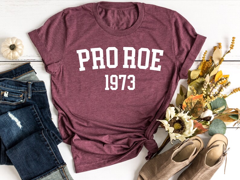 May include: A maroon t-shirt with white text that reads "PRO ROE 1973". The shirt is laid flat on a white wooden surface with a pair of blue jeans, a small white pumpkin, and a bouquet of dried flowers.