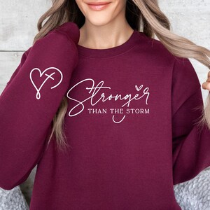 Pu&ograve; includere: Felpa color borgogna con testo bianco che recita "Stronger than the storm" e un cuore con una croce all'interno sulla manica.