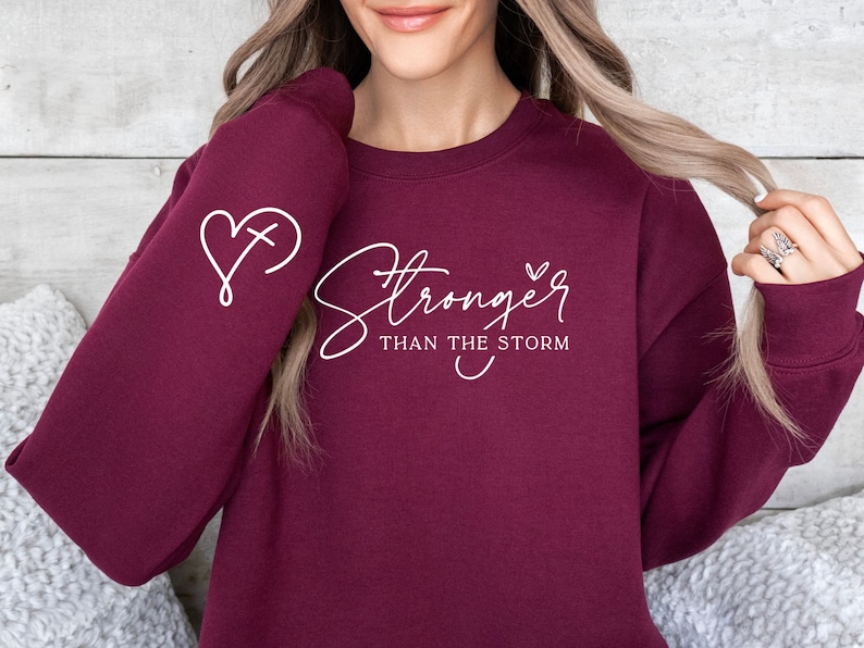 Pu&ograve; includere: Felpa color borgogna con testo bianco che recita "Stronger than the storm" e un cuore con una croce all'interno sulla manica.