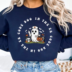 May include: A navy blue sweatshirt with a white graphic of a ghost wearing a nurse's hat and holding a syringe and a pumpkin. The text "Boo in the ER Code Boo in the ER Code" is printed in a circle around the ghost.
