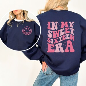 May include: Navy blue sweatshirt with a pink graphic on the back that says "In My Sweet Sixteen Era" and a pink smiley face with a heart and lightning bolt on the front.