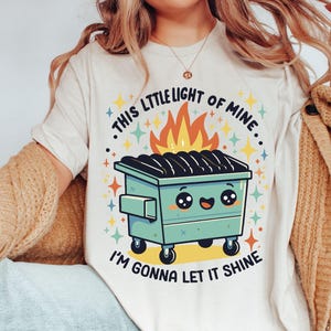 May include: White t-shirt with a graphic of a green dumpster on fire with a smiling face and the text "This little light of mine, I'm gonna let it shine."