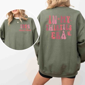 May include: A green sweatshirt with a pink and red graphic on the back that says "In My Thirties Era". The front of the sweatshirt has a pink and red graphic that says "1991" with the text "Golden Era" underneath.