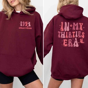 May include: Burgundy hoodie with a graphic on the back that says "In My Thirties Era" in a retro font with stars and a butterfly. The front of the hoodie has a graphic that says "1994 Amelia's Version".