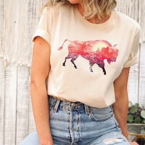 May include: A cream colored t-shirt with a graphic of a silhouette of a bison with a sunset landscape inside the silhouette.