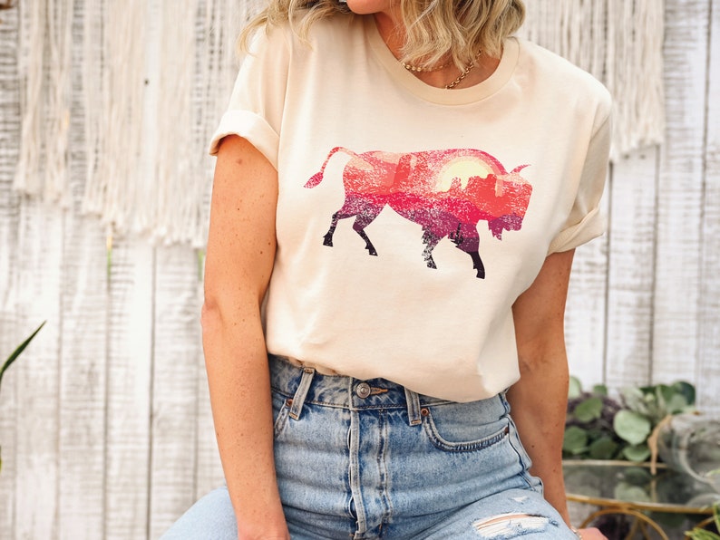 May include: A cream colored t-shirt with a graphic of a silhouette of a bison with a sunset landscape inside the silhouette.