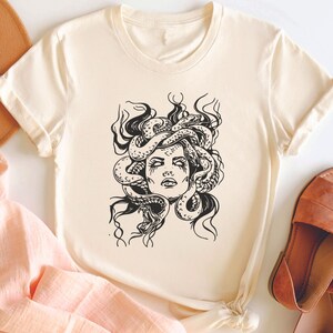 May include: A cream-colored t-shirt with a black and white illustration of Medusa, a mythical Greek figure with snakes for hair.