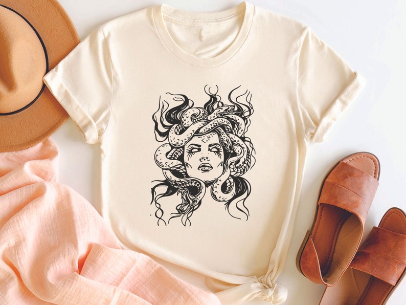 May include: A cream-colored t-shirt with a black and white illustration of Medusa, a mythical Greek figure with snakes for hair.