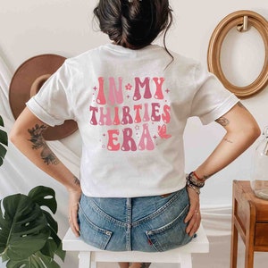 May include: White t-shirt with pink text that reads "In My Thirties Era" with stars and a butterfly graphic.