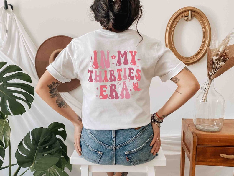 May include: White t-shirt with pink text that reads "In My Thirties Era" with stars and a butterfly graphic.