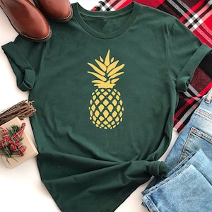 May include: A dark green t-shirt with a yellow pineapple graphic. The shirt is folded and laid on a white surface with a plaid fabric and blue jeans.