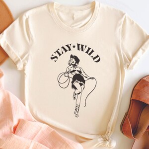 May include: A cream colored t-shirt with a black graphic of a woman in a cowboy hat holding a lasso. The text "STAY WILD" is printed above the graphic.