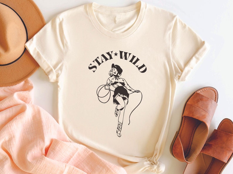 May include: A cream colored t-shirt with a black graphic of a woman in a cowboy hat holding a lasso. The text "STAY WILD" is printed above the graphic.