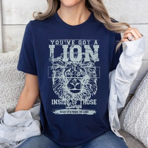 May include: Navy blue t-shirt with a distressed white graphic of a lion's head and the text "You've got a lion inside of those lungs. So get up & praise the Lord."
