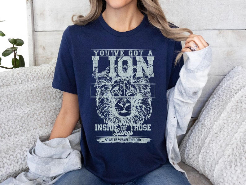 May include: Navy blue t-shirt with a distressed white graphic of a lion's head and the text "You've got a lion inside of those lungs. So get up & praise the Lord."
