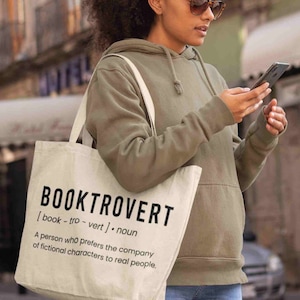 May include: Off-white tote bag with the word "BOOKTROVERT" in large black letters. The definition of the word is printed below. The person in the image is wearing a green hoodie and sunglasses.