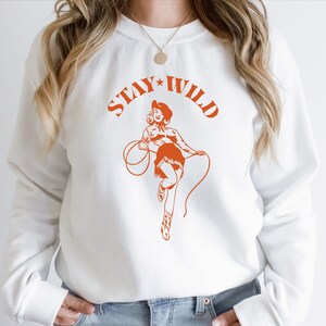 May include: White sweatshirt with a graphic of a woman in a cowboy hat and the text "Stay Wild" in orange.