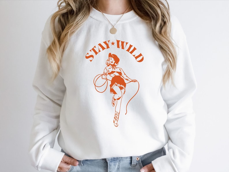 May include: White sweatshirt with a graphic of a woman in a cowboy hat and the text "Stay Wild" in orange.