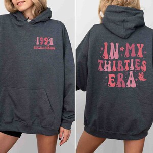 May include: A dark gray hooded sweatshirt with a pink graphic on the back that says "IN MY THIRTIES ERA" with stars and a butterfly. The front of the sweatshirt has a pink graphic that says "1994 AMELIA'S VERSION".