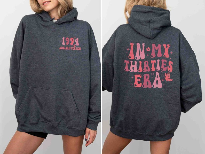 May include: A dark gray hooded sweatshirt with a pink graphic on the back that says "IN MY THIRTIES ERA" with stars and a butterfly. The front of the sweatshirt has a pink graphic that says "1994 AMELIA'S VERSION".