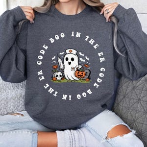 May include: A dark gray sweatshirt with a white graphic of a ghost wearing a nurse's hat and holding a pumpkin. The text around the ghost reads "Boo in the ER Code the Boo in the ER Code".
