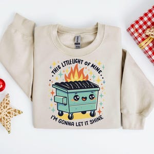 May include: A beige sweatshirt with a graphic of a green dumpster with flames coming out of it. The text on the sweatshirt reads "This little light of mine... I'm gonna let it shine."