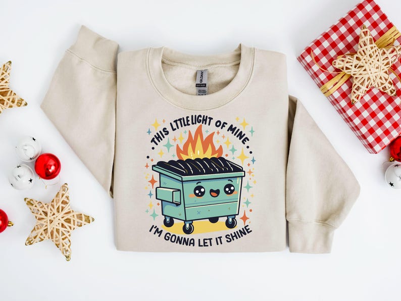 May include: A beige sweatshirt with a graphic of a green dumpster with flames coming out of it. The text on the sweatshirt reads "This little light of mine... I'm gonna let it shine."