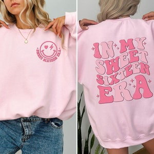 May include: Pink sweatshirt with a smiley face with a heart and lightning bolt on the front and the text "In My Sweet Sixteen Era" on the back.