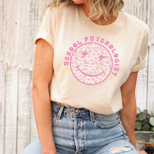 May include: A cream colored t-shirt with a pink and white smiley face graphic that says "School Psychologist".